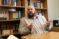 A man sits in front of a bookshelf in his office, and speaks to someone while gesturing with his hands