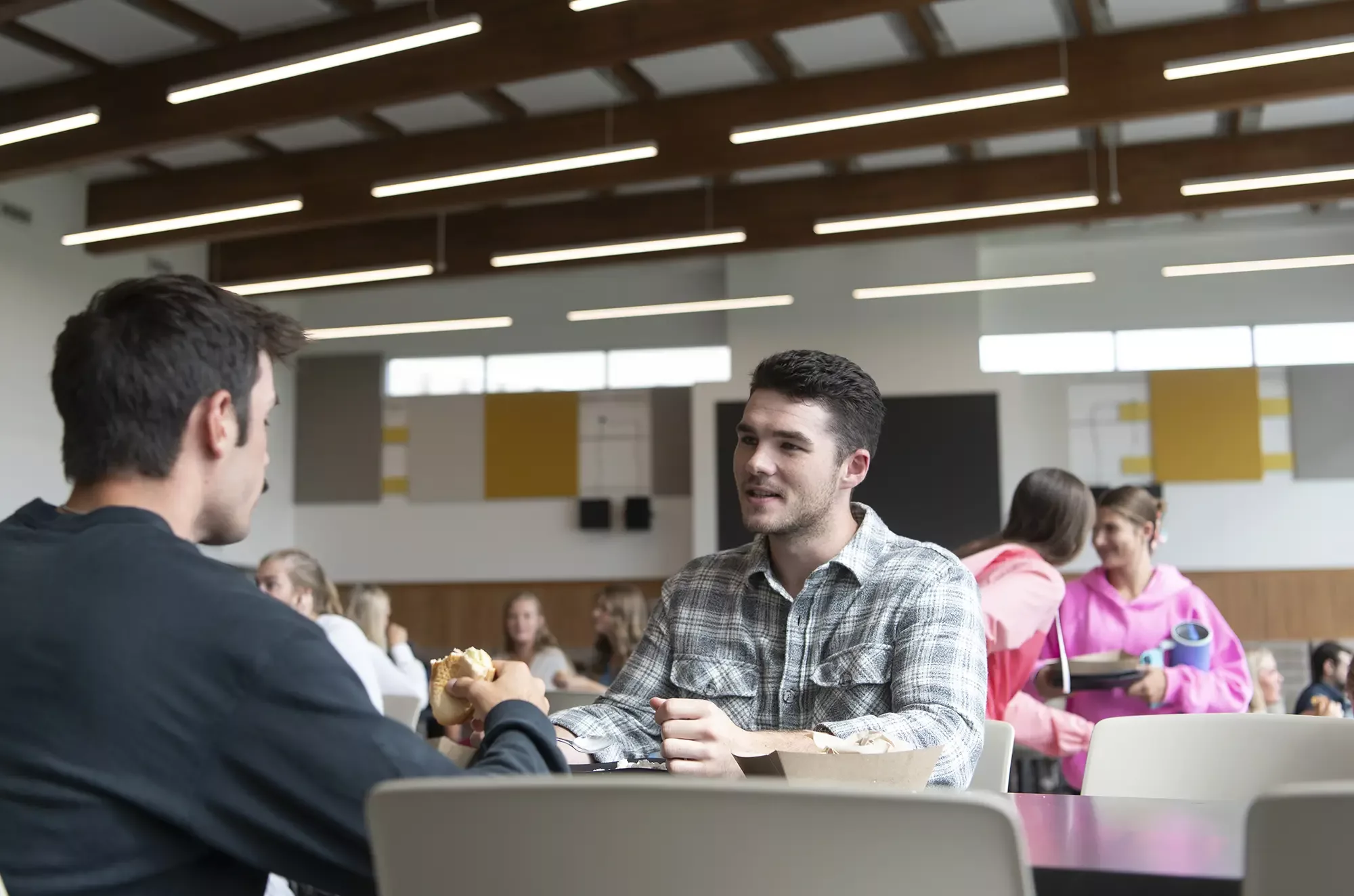Young man at table converses with another young man in large space with expansive wood beams on ceiling