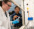 A Dordt student in a lab coat and safety glasses holding a chemical vase while her Professor points at the vase and explains something