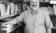 Black and white photo of bearded man standing in corner of room amidst bookshelves