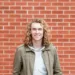 A picture of a man with long curly hair standing in front of a brick wall.